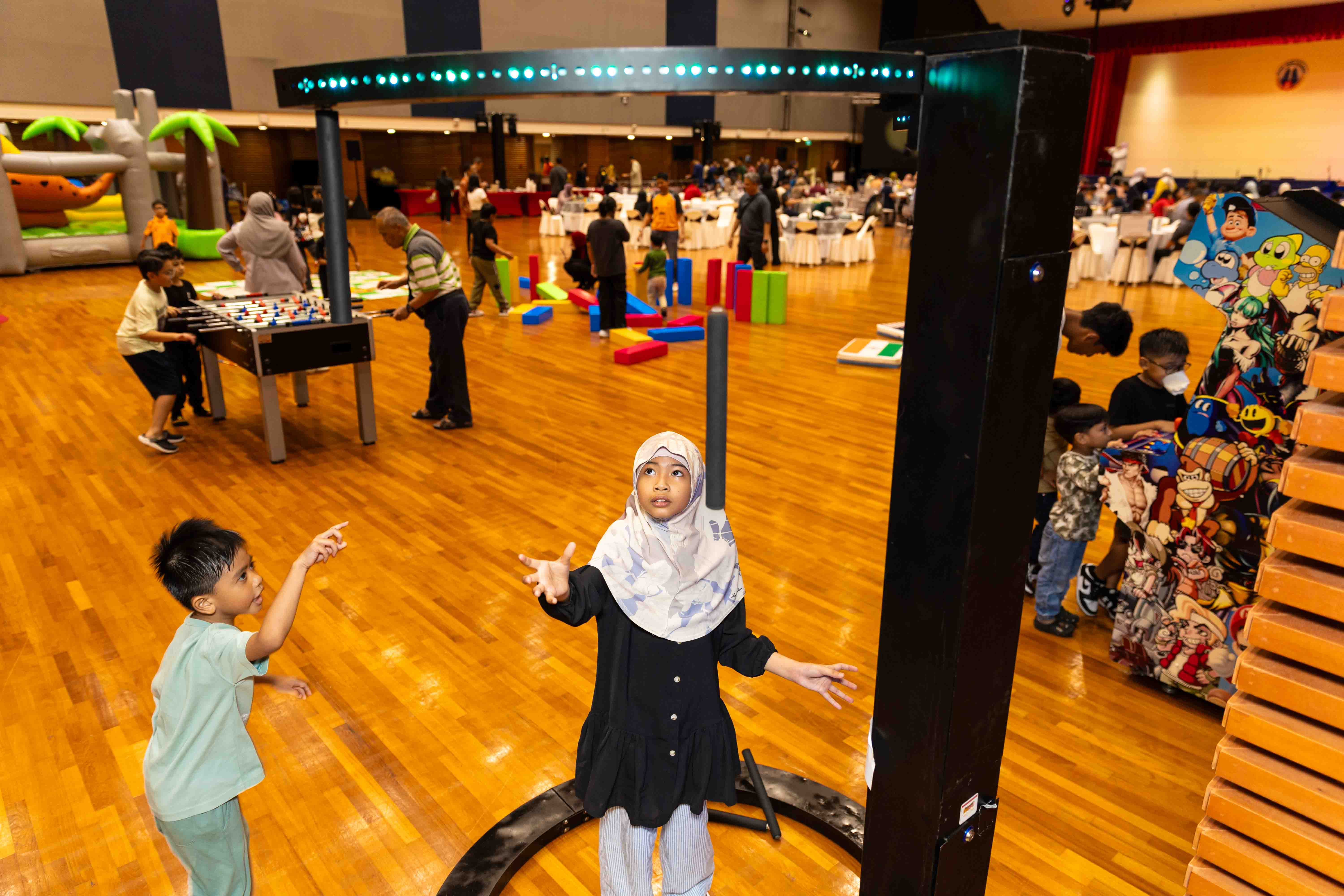 Two children reach for falling sticks in a reaction game while other families enjoy arcade, table football, inflatable play equipment and giant block games in the hall behind them.